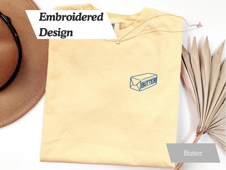 May include: A light yellow t-shirt with an embroidered butter design. The design is a blue outline of a butter package with the word "Butter" inside. The shirt also has the words "Embroidered Design" in a white banner.
