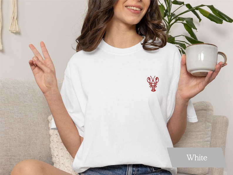 Woman wearing a white Comfort Colors t-shirt with minimalist embroidered lobster design, coastal graphic tee perfect for tomato girl summer and beach-inspired outfits