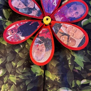 May include: A red plastic pinwheel featuring six petal-shaped blades, each displaying a unique photograph with a holographic glitter finish. The blades are attached to a central yellow hub, and the entire structure is mounted on a thin black rod.