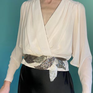 Puede incluir: Blusa color crema con cuello en V y mangas largas. La blusa se ciñe a la cintura con un cinturón decorativo con un diseño de cuentas en tonos plateados, marrones y blancos. La modelo lleva una falda negra.