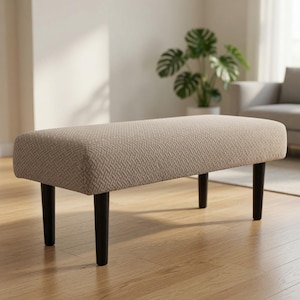 Puede incluir: Un banco rectangular con tapicería de tela beige y un patrón tejido texturizado. El banco se apoya sobre cuatro patas rectas y cilíndricas de color marrón oscuro, lo que proporciona una base estable para el asiento acolchado.