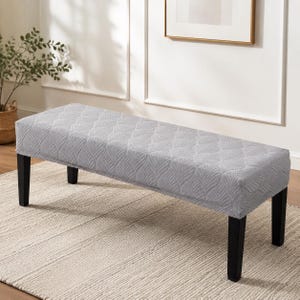 Puede incluir: Un banco rectangular con una funda elástica de color gris claro que presenta un patrón de hojas repetitivo y texturizado. El banco se apoya sobre cuatro patas rectas de madera negra maciza, proporcionando una base estable.