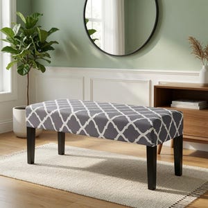 May include: A rectangular bench featuring a dark grey upholstered seat with a white quatrefoil pattern. The bench is supported by four straight, dark brown wooden legs. The seat is flat and padded, providing a stable surface for seating.