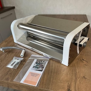 Könnte beinhalten: Ein weißer und Edelstahl-Teigausroller für den Hausgebrauch. Der Ausroller hat eine große Walze, einen Griff und eine transparente Plastiktüte mit einer Anleitung. Der Ausroller steht auf einem Holztisch.