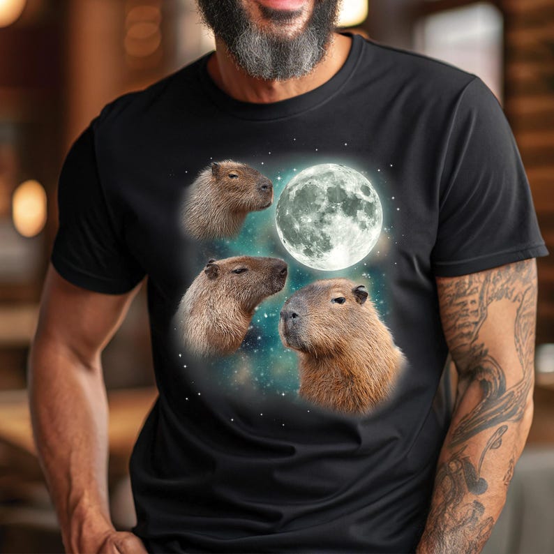 May include: Black t-shirt featuring a graphic of capybaras and a full moon against a starry background. The capybaras are depicted in shades of brown, with the moon in white and gray.