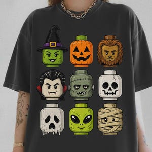May include: A charcoal gray t-shirt featuring a graphic print of nine stylized block-style character heads arranged in a grid. The characters include a witch, a jack-o'-lantern, a werewolf, a vampire, a monster, a skull, a ghost, an alien, and a mummy.
