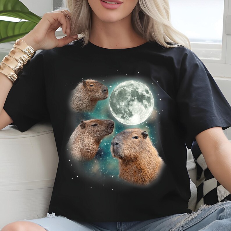 May include: Black t-shirt featuring a graphic design of three capybaras and a full moon against a starry background. The capybaras are depicted in shades of brown, and the moon is a bright white. The design is centered on the front of the shirt.