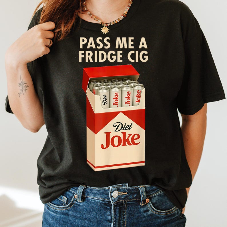 May include: Black t-shirt with the slogan "PASS ME A FRIDGE CIG". The shirt displays a graphic of a cigarette box labelled "Diet Joke" filled with soda cans. The cans are silver with the word "Joke" printed on them.