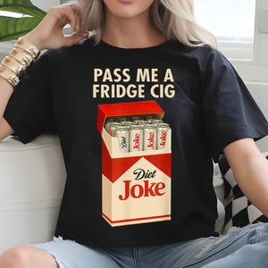 May include: Black t-shirt featuring the text "PASS ME A FRIDGE CIG" above a graphic of a cigarette pack. The pack is red and white, with "Diet Joke" printed on it, and contains silver cans labeled "Diet Joke".