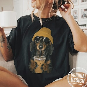 Puede incluir: Una camiseta negra con un estampado gráfico de un perro salchicha que lleva un gorro de color amarillo mostaza y gafas de sol redondas negras, sosteniendo un vaso de plástico transparente con pajita lleno de café helado.