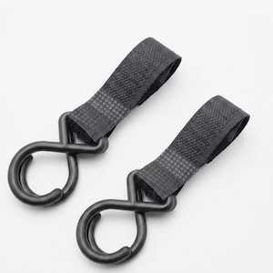 May include: Two black, heavy-duty bungee cord hooks with woven nylon straps. The hooks are a matte black finish and have a distinctive looped design. The straps are a dark gray color and appear to be durable. The hooks are likely used for securing items.