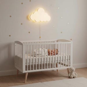 Könnte beinhalten: Ein weißes Babybett mit einem schlafenden Baby, einer wolkenförmigen Wandleuchte und einem Teddybären. Das Babybett hat ein Lattenrost-Design und Holzbeine. Die Wand ist mit goldenen Sternaufklebern dekoriert.