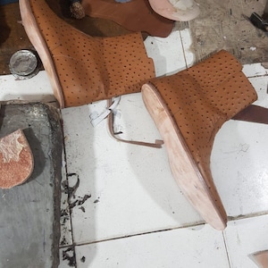 Pu&ograve; includere: Un paio di stivaletti in pelle color cuoio con un motivo traforato su tutta la superficie superiore. Gli stivaletti presentano suole chiare non rifinite e sono mostrati in un laboratorio con vari ritagli di pelle e attrezzi visibili nelle vicinanze.