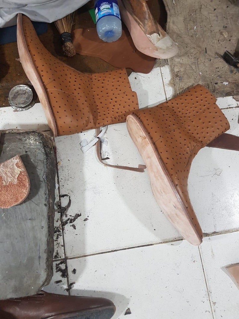 Pu&ograve; includere: Un paio di stivaletti in pelle color cuoio con un motivo traforato su tutta la superficie superiore. Gli stivaletti presentano suole chiare non rifinite e sono mostrati in un laboratorio con vari ritagli di pelle e attrezzi visibili nelle vicinanze.