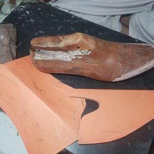 Pu&ograve; includere: Una forma per scarpe in legno marrone con superficie usurata e un piccolo foro metallico, posizionata accanto a un pezzo di cartamodello sintetico piatto color pesca con linee disegnate a mano per la creazione di calzature.
