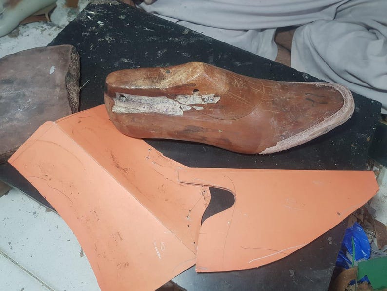 Pu&ograve; includere: Una forma per scarpe in legno marrone con superficie usurata e un piccolo foro metallico, posizionata accanto a un pezzo di cartamodello sintetico piatto color pesca con linee disegnate a mano per la creazione di calzature.