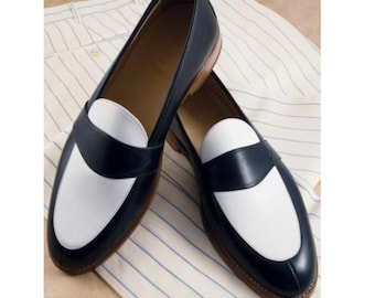 May include: A pair of two-tone loafers featuring a dark navy blue leather exterior with a contrasting white leather vamp panel. The shoes have a classic slip-on design with a rounded toe, a low heel, and a tan interior lining with visible stitching.