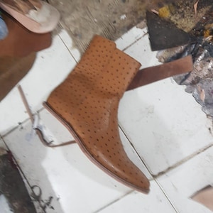 Pu&ograve; includere: Uno stivaletto color cuoio caratterizzato da una distinta texture puntinata su tutta la superficie. Lo stivale presenta una punta affusolata, un gambale alto e una suola liscia e piatta. Il materiale mostra un colore marrone chiaro uniforme.