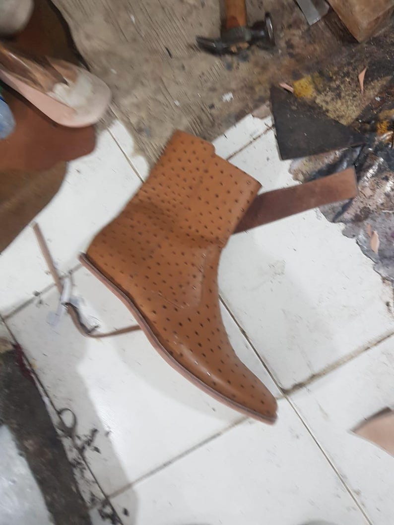 Pu&ograve; includere: Uno stivaletto color cuoio caratterizzato da una distinta texture puntinata su tutta la superficie. Lo stivale presenta una punta affusolata, un gambale alto e una suola liscia e piatta. Il materiale mostra un colore marrone chiaro uniforme.