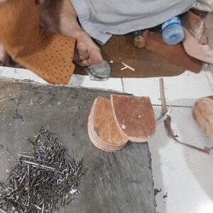 Pu&ograve; includere: Un pezzo di pelle color cuoio con una trama punteggiata, due componenti per tacco in pelle marrone impilati e un mucchio di piccoli chiodi metallici su una superficie di lavoro grigia. Il pezzo di pelle &egrave; sollevato mentre i tacchi e i chiodi restano.