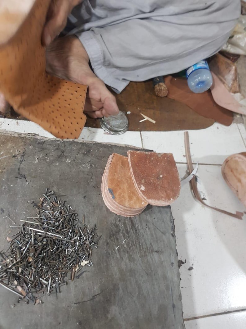 Pu&ograve; includere: Un pezzo di pelle color cuoio con una trama punteggiata, due componenti per tacco in pelle marrone impilati e un mucchio di piccoli chiodi metallici su una superficie di lavoro grigia. Il pezzo di pelle &egrave; sollevato mentre i tacchi e i chiodi restano.