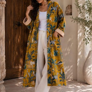 Op de afbeelding: Een lange, mosterdgele dusterjas met een levendige jungleprint van zebra's, luipaarden en palmbomen. De jas heeft lange mouwen met wit en beige gestreepte boorden, een open voorzijde en twee zijzakken, gedragen over een wit hemdje en broek.