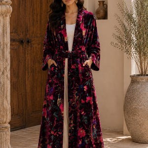 May include: A long, open-front velvet robe in a deep burgundy hue, featuring a vibrant floral pattern with pink, blue, and green botanical motifs. The garment includes long sleeves, a matching tie belt at the waist, and two front pockets.