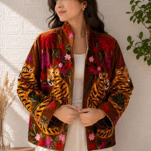 Può includere: Una giacca in velluto marrone con ricami colorati di tigri e motivi floreali. La giacca presenta un colletto alto, maniche lunghe e un design aperto sul davanti, con dettagli ricamati nelle tonalità del rosso, rosa e giallo.