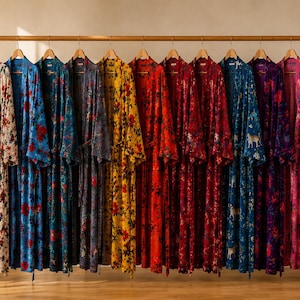 Op de afbeelding: Een rij van twaalf kamerjassen met lange mouwen en vloerlengte, hangend aan een houten rek. De kamerjassen zijn voorzien van diverse levendige bloemen- en dierenpatronen in kleuren zoals wit, blauw, geel, rood, bordeaux, paars, roze en donkergroen.