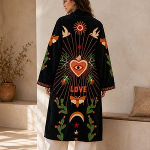 May include: A black long-line jacket featuring colorful embroidery on the back. The design includes a central heart with an eye, a sun, birds, moths, cacti, and the word LOVE. The jacket has long sleeves and is paired with loose white trousers.