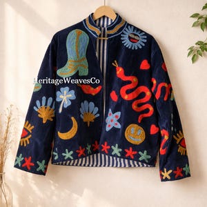 Puede incluir: Chaqueta azul marino con diseños bordados coloridos, incluyendo una bota de vaquero, una serpiente, un sol y flores. La chaqueta tiene un forro a rayas y está colgada de una percha de madera. El texto "Heritage WeavesCo" es visible.