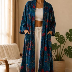 May include: A long, open-front robe in teal blue velvet featuring a vibrant floral and bird pattern with red and pink blossoms and green foliage. The garment includes long sleeves, a shawl collar, and two front patch pockets, worn over a white tank top.