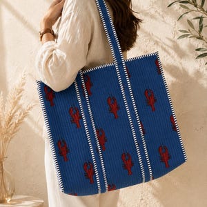 Könnte beinhalten: Eine große, rechteckige blaue Tragetasche mit einem sich wiederholenden Muster aus roten Hummern. Die Tasche besteht aus gestepptem Stoff und verfügt über zwei Schulterriemen mit weiß-dunkelblau gestreifter Verzierung an den Kanten.