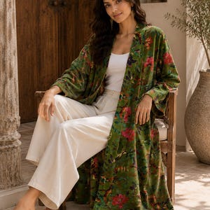 May include: A long-sleeved, open-front velvet duster robe in an olive green hue, featuring a vibrant floral pattern with pink, red, and purple blossoms. The garment is worn over a plain white tank top and loose-fitting, wide-leg cream-coloured trousers.
