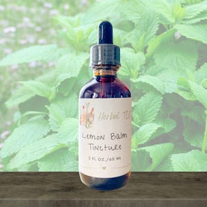 May include: A clear glass bottle with a dark liquid and a dropper. The label reads "Lemon Balm Tincture" and "2 fl oz / 60 ml". The background features green foliage, suggesting a natural product.