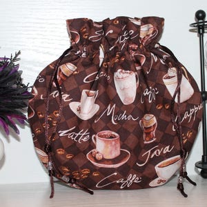Puede incluir: Una bolsa marrón con cordón y un estampado de café. La bolsa presenta imágenes de tazas de café, granos y bebidas como latte, moca y capuchino. El fondo es un patrón de cuadros. El cordón es marrón con cuentas decorativas.