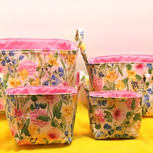 May include: Four fabric storage baskets with floral patterns. The baskets have a pink interior and handles. The floral pattern includes pink, yellow, and blue flowers with green leaves. The baskets are arranged in a nested configuration.