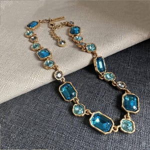 May include: A gold-toned necklace featuring a series of blue and teal-colored rectangular and square-shaped gemstones. The necklace is laid on a neutral-colored fabric, showcasing its design and color variations.