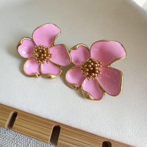 May include: A pair of pink flower-shaped earrings with gold-colored trim and a textured center. The earrings are set against a white background.
