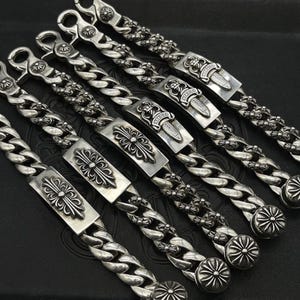 Pode incluir: Pulseiras de corrente prateadas com desenhos ornamentados de cruzes e adagas. Cada pulseira apresenta uma corrente grossa e um fecho decorativo. As pulseiras são exibidas em uma superfície escura.