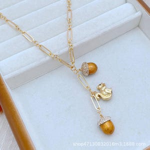 Peut inclure: Collier doré avec un pendentif en forme d'écureuil et de glands. La chaîne délicate est composée de maillons rectangulaires. Les glands ont des corps bruns et des chapeaux incrustés de cristaux. Un petit écureuil doré est positionné entre les glands.