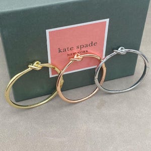 May include: Three knot-shaped bangles in gold, rose gold, and silver. The bracelets are displayed in front of a green box with a pink label that reads "kate spade NEW YORK". The bangles are on a light grey surface.