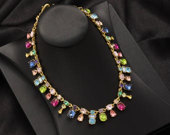 Può includere: Una collana dorata con una catena delicata e una serie di pietre preziose colorate. La collana presenta gemme di varie forme e dimensioni in tonalità rosa, blu, verde e giallo. È esposta su un espositore per gioielli nero.