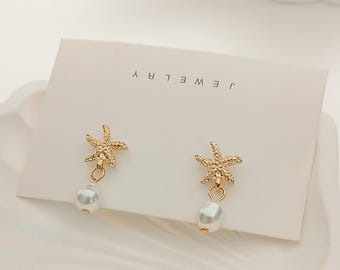 May include: Gold-colored starfish earrings with a dangling white pearl. The earrings are displayed on a white card with the word "JEWELRY" printed on it. The earrings are on a white surface.