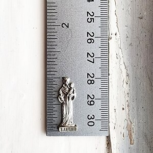 May include: A silver charm featuring a depiction of Saint Anthony holding a baby. The charm is approximately 1 inch tall and has the words "S. ANTONIO" engraved on the bottom.