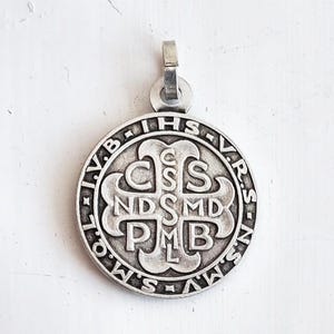 May include: A silver-colored pendant with a circular design. The pendant features embossed lettering and symbols, including the letters "IHS", "CSS", "NDSMD", "PMB", and others. It has a small loop at the top for a chain.