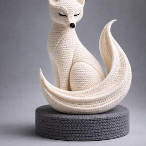 Op de afbeelding: Een crèmekleurig, gehaakt vossenbeeldje met gesloten ogen en een zwarte neus. De vos zit op een grijze, ronde, gehaakte basis. De staart van de vos krult om zijn lichaam. De achtergrond is effen grijs.