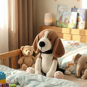 Pode incluir: Uma cena aconchegante de quarto infantil com um grande cão de crochê feito à mão com detalhes marrons e brancos, sentado em uma cama com um edredom estampado em azul claro. Um ursinho de pelúcia e outros brinquedos também estão presentes.