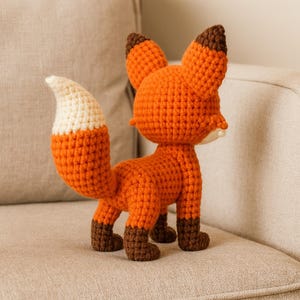 K&ouml;nnte beinhalten: Ein geh&auml;keltes Fuchspielzeug in Orange, Braun und Creme. Der Fuchs hat einen flauschigen, wei&szlig;spitzigen Schwanz, braune Pfoten und braunspitzige Ohren. Das handgefertigte Stofftier steht auf einer neutralfarbenen Oberfl&auml;che.