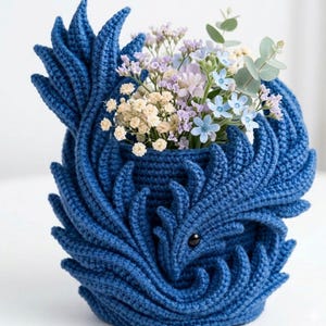May include: A handcrafted, blue crochet vase with a unique, swirling design. The vase is filled with a variety of colorful flowers, including light blue, purple, and white blooms, and green foliage. The vase has a black eye detail.
